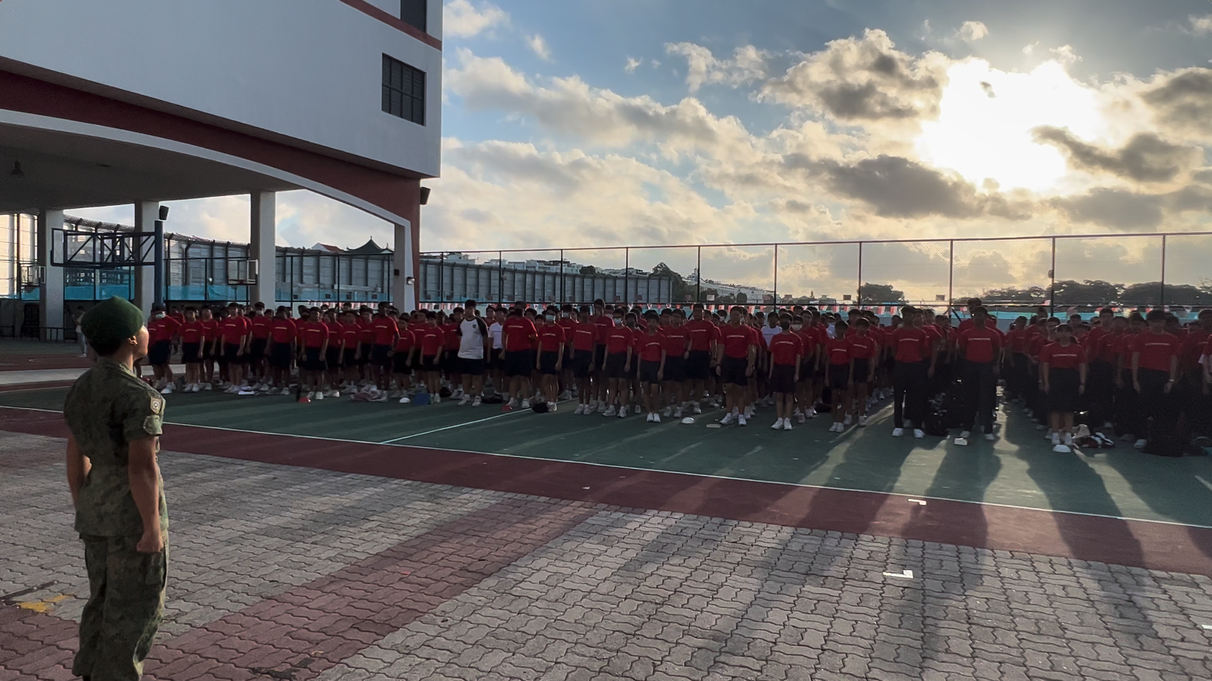 A soldier in camouflage faces a large group of students in red shirts standing in formation on an outdoor court.